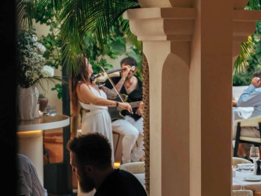 An outdoor dining area with a covered section, palm trees, and a person playing a guitar.