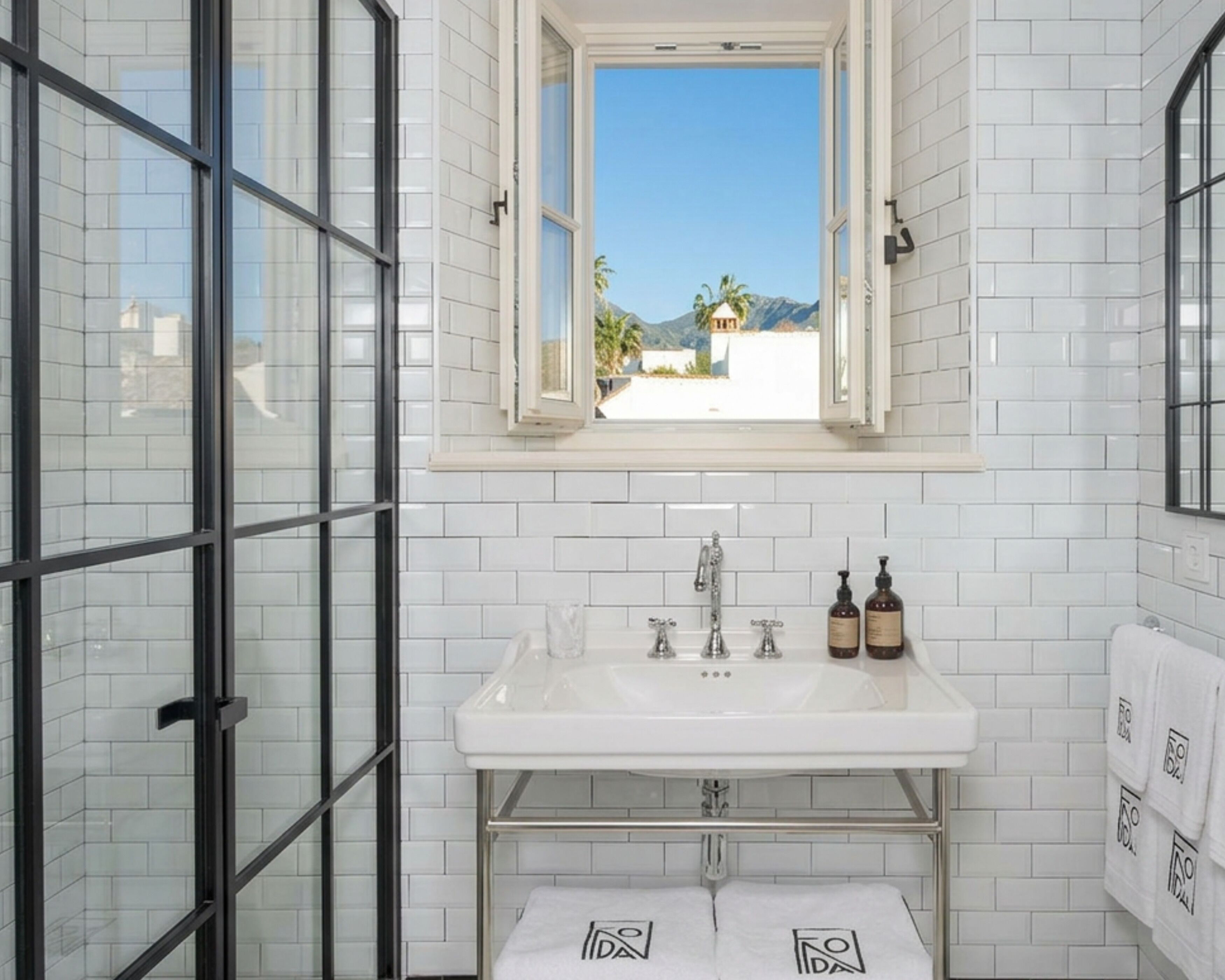 A bathroom with a white tiled wall, a window with a view of palm trees and mountains, a white sink with two bottles of soap, and a towel rack with towels.