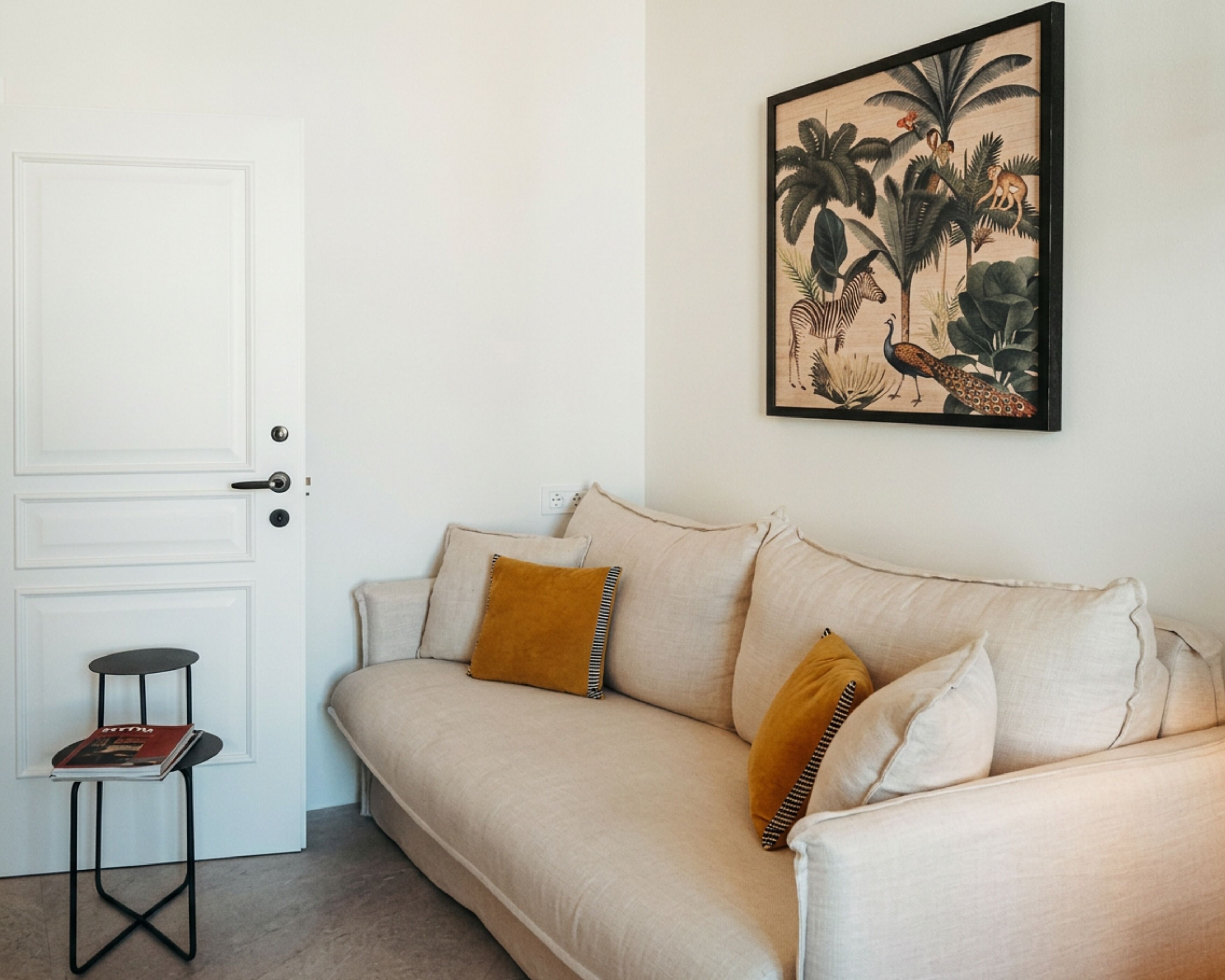 A living room with a beige sofa, two cushions, a small table with a magazine, and a framed picture on the wall.
