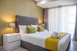 A neatly made bed with white and yellow bedding, green pillows, and a yellow throw blanket. There are two bedside lamps on white nightstands. - Maison Moz - Polana Plaza VII (Maputo)