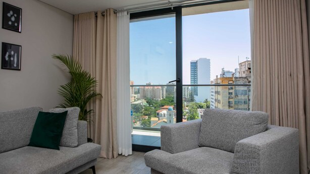 A modern living room with a grey sofa, a green pillow, and a large window with curtains. - Maison Moz - Polana Plaza IV (Maputo)