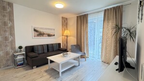 A modern living room with a sofa, a coffee table, a lamp, a TV, and a window with curtains. - Vacation apartment Nara (Esens)