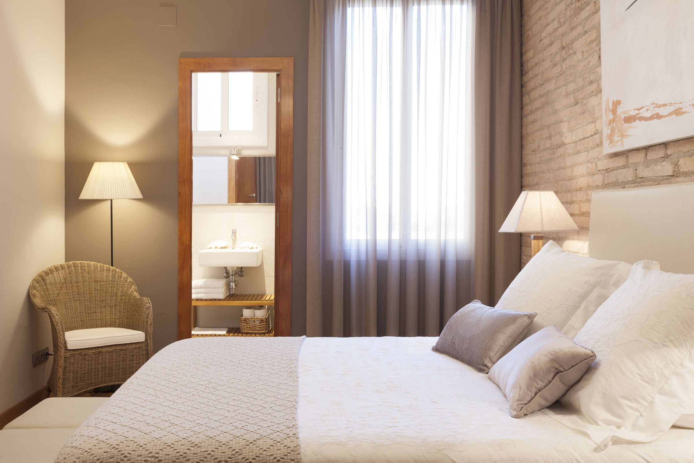 A hotel room with a bed, two lamps, a wicker chair, a mirror, and a window with curtains.