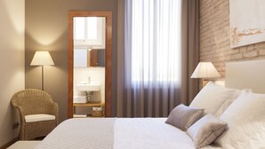 Una habitación de hotel con una cama, dos lámparas, un sillón de mimbre, un espejo y una ventana con cortinas.