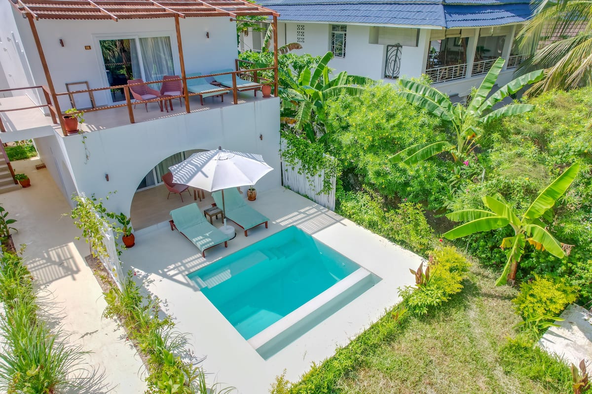 Una casa de dos pisos con piscina, mobiliario exterior y abundante vegetación.