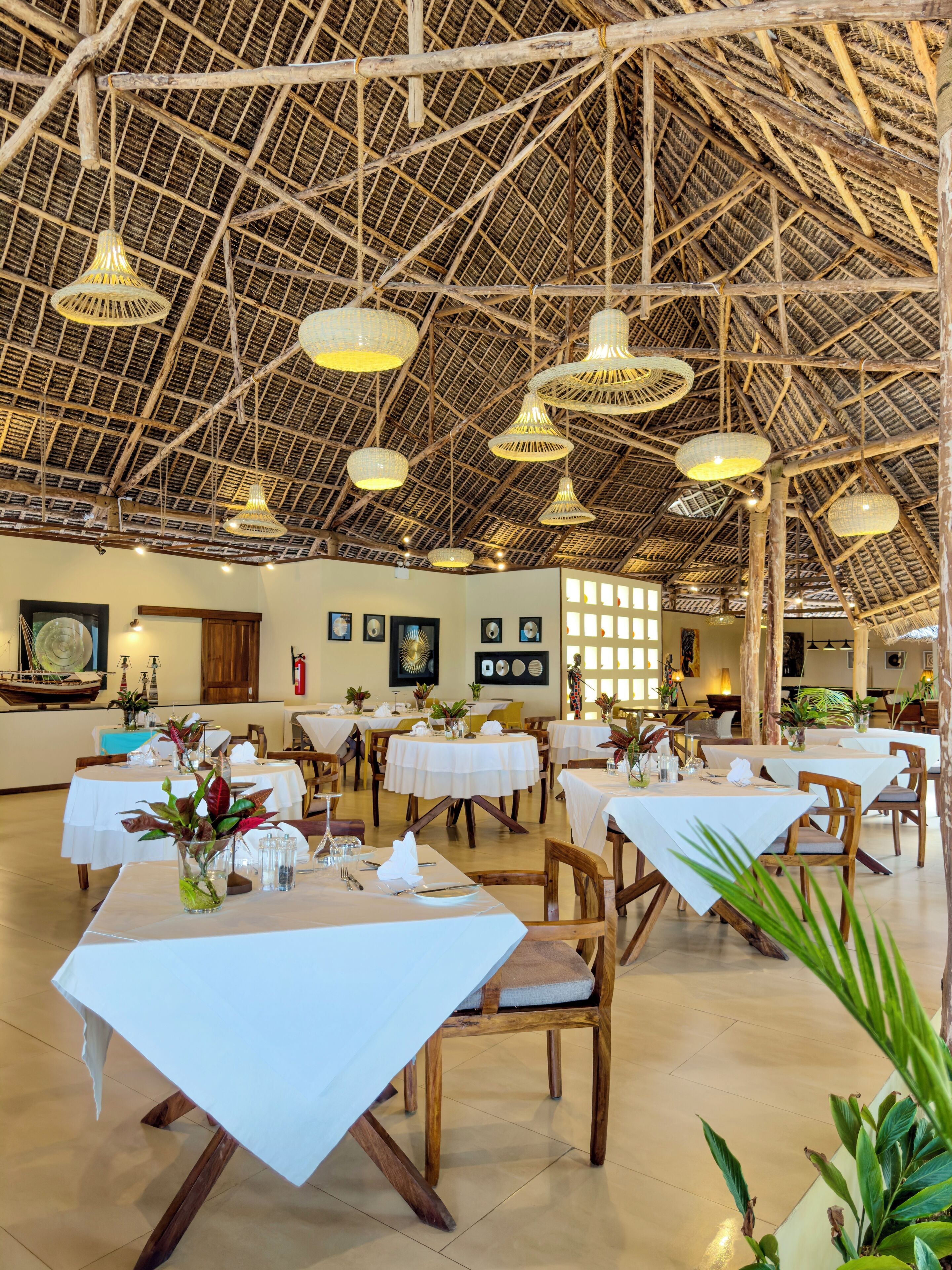 A restaurant with high ceilings, wooden beams, and hanging wicker lamps. Tables are set with white tablecloths and centerpieces.