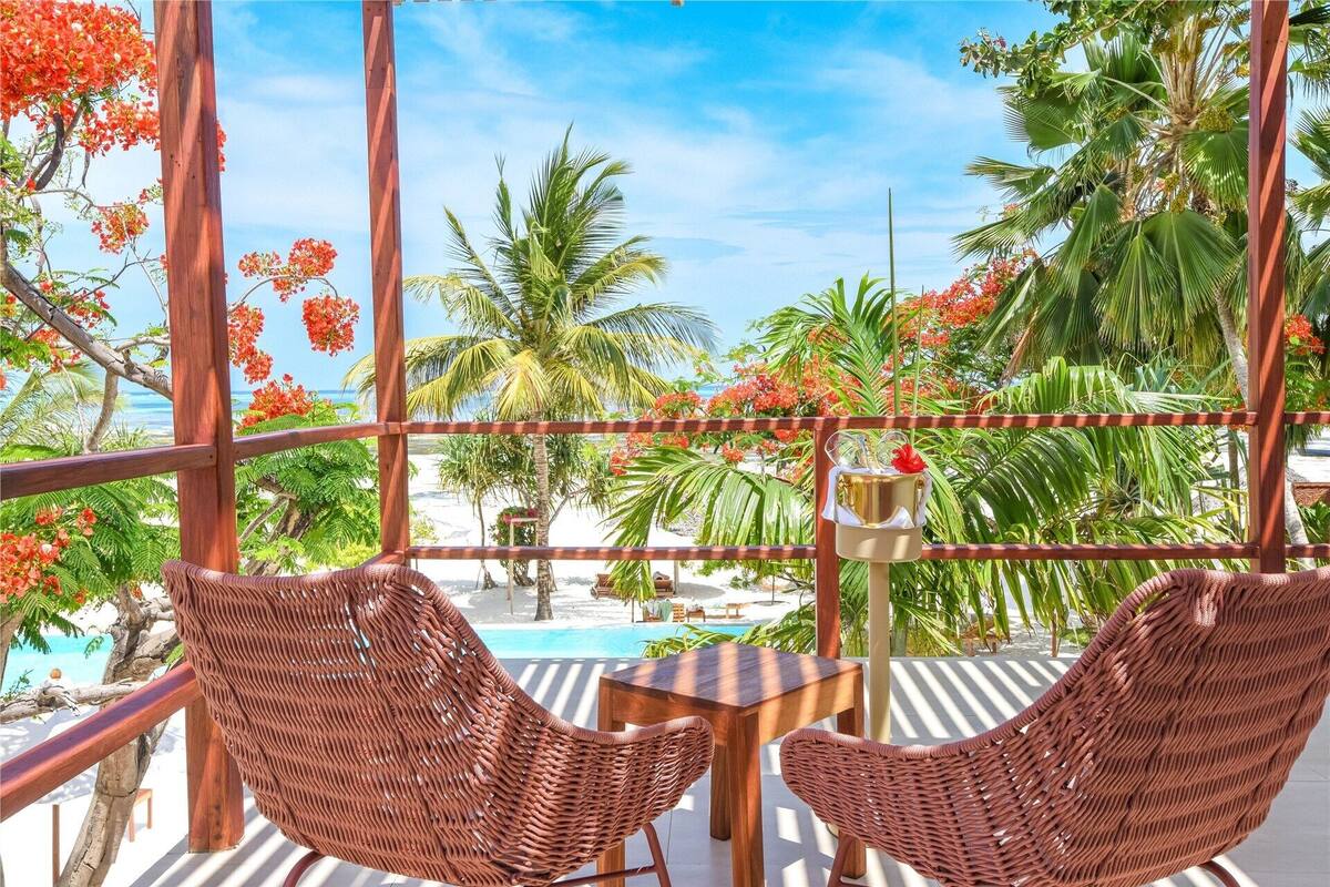 Un balcón con sillas de mimbre, una mesa y vistas a una piscina y palmeras.
