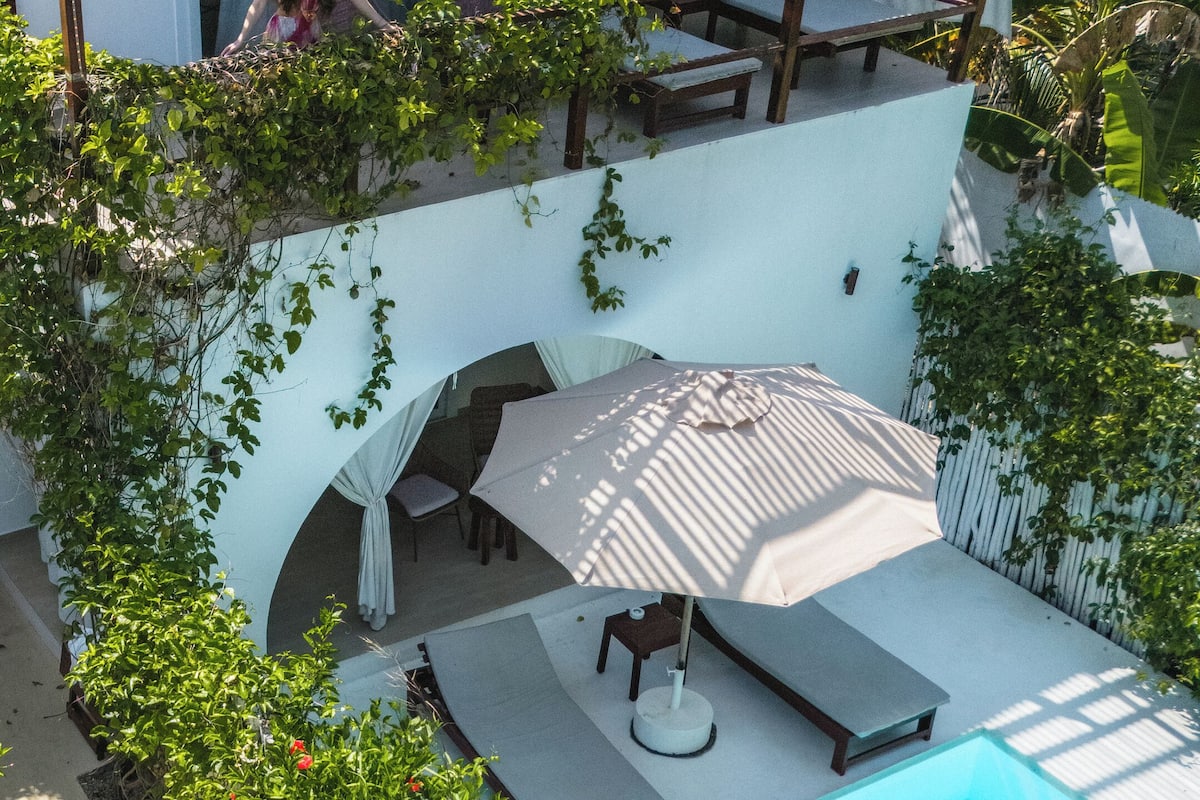 Una piscina con sombrilla, rodeada de exuberante vegetación y un patio con mobiliario exterior.