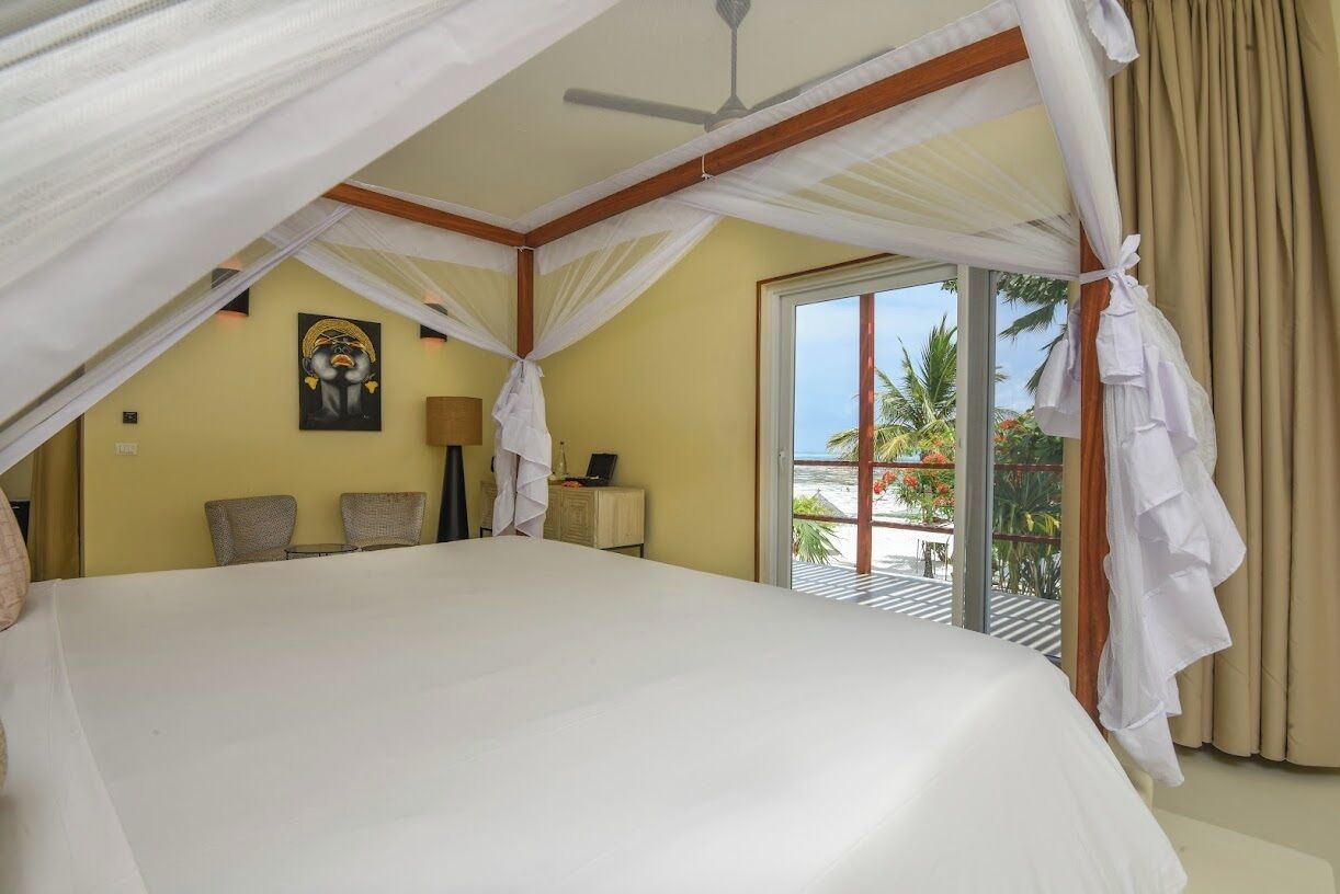 A large bed with a canopy, a painting on the wall, a desk with a lamp, and a view of palm trees through a glass door.