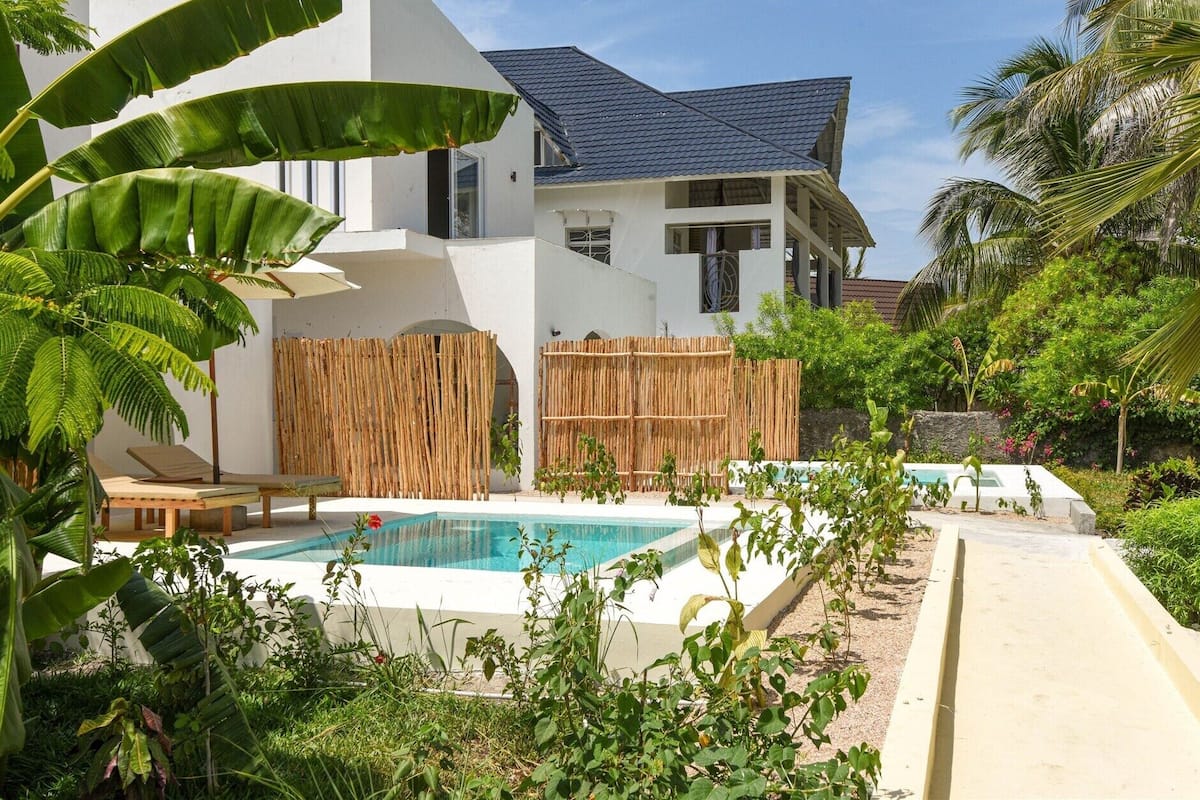 Una piscina con valla de bambú, un edificio blanco con techo oscuro y vegetación tropical.