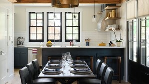 A modern kitchen with a dining area, featuring a large wooden table set for six, a hanging light fixture, and a view of the outside through large windows.