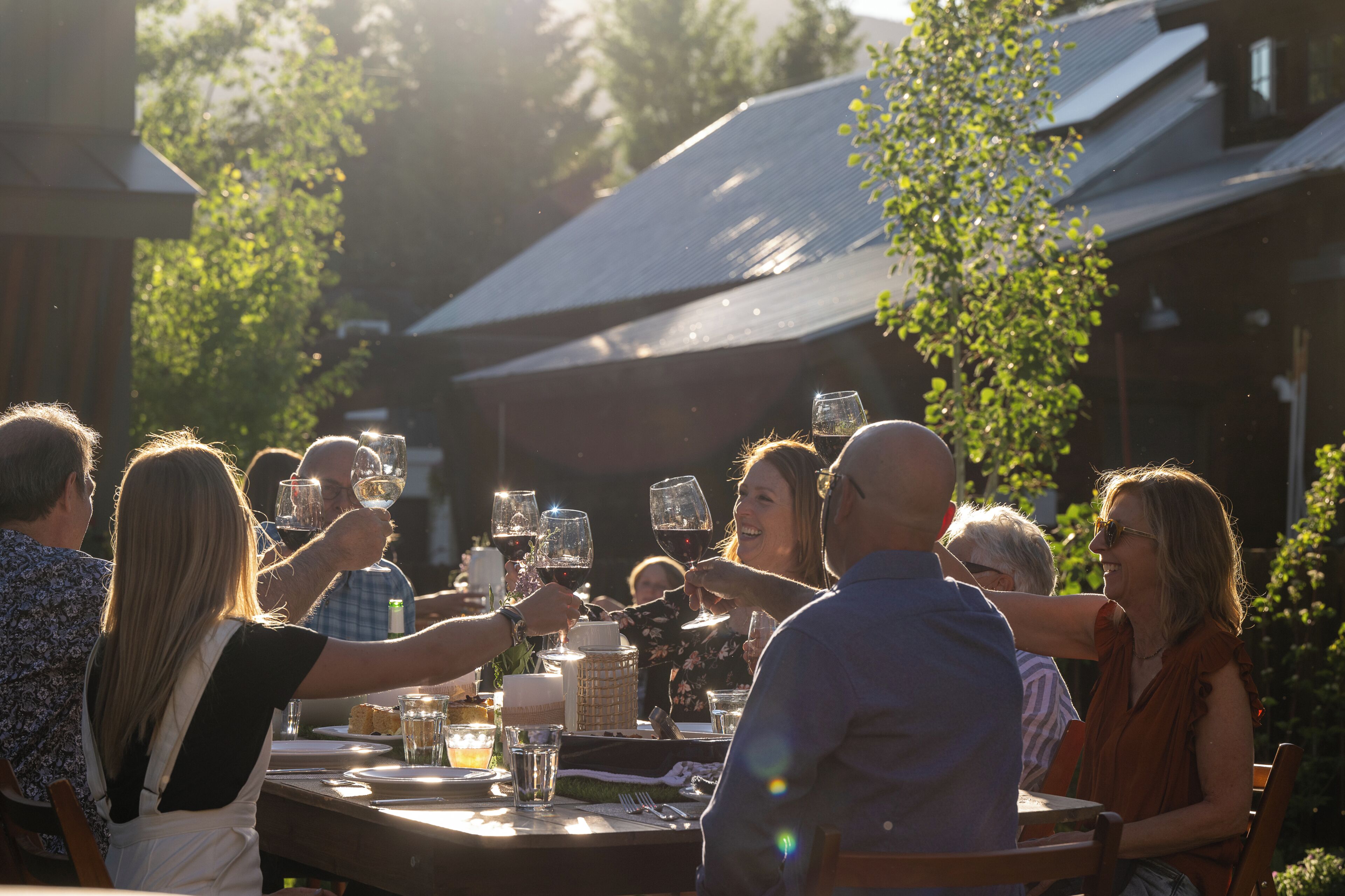 A group of people toasting at an outdoor table with wine glasses, surrounded by trees and buildings.