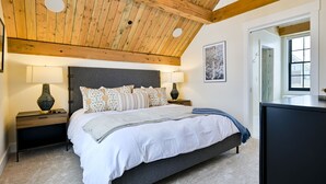 A bedroom with a wooden ceiling, a bed with patterned pillows, two bedside lamps, and a large window. - Academy Place | Bluebell (Crested Butte)
