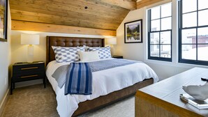 A bedroom with a wooden ceiling, a large bed with a checkered headboard, two bedside tables, and a window with a view of buildings. - Academy Place | Fairy Slipper (Crested Butte)