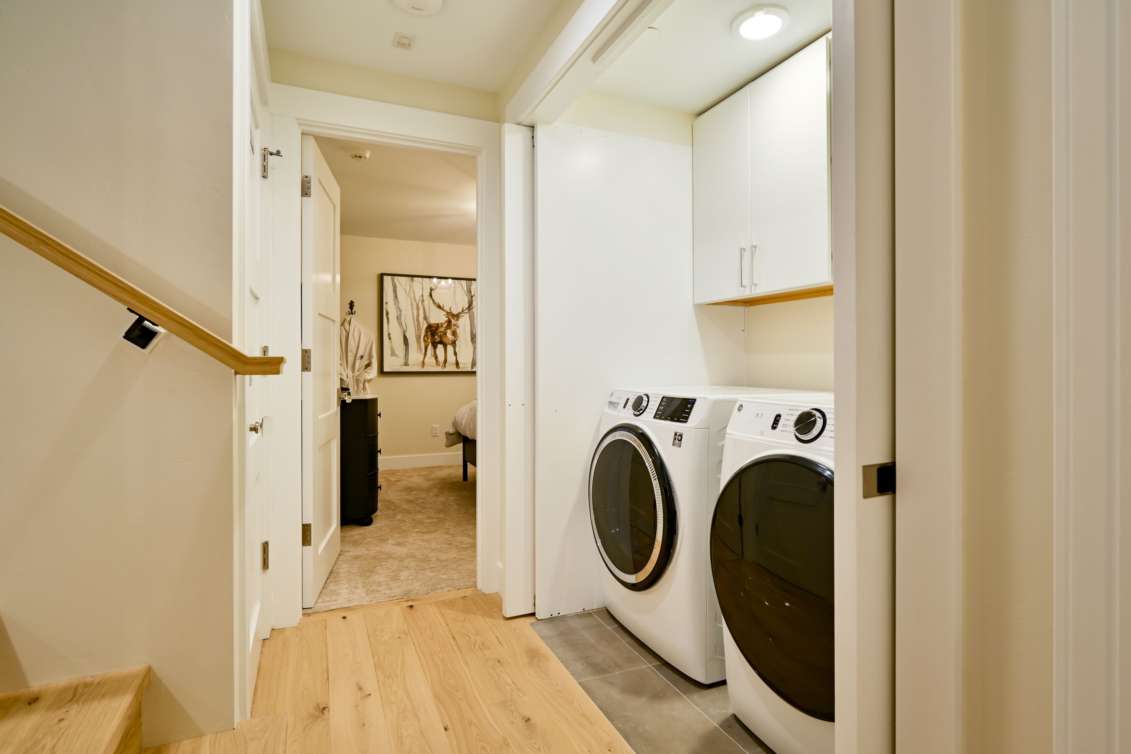 A laundry room with two front-loading washing machines, a wooden floor, and a hallway leading to another room with a framed picture on the wall.