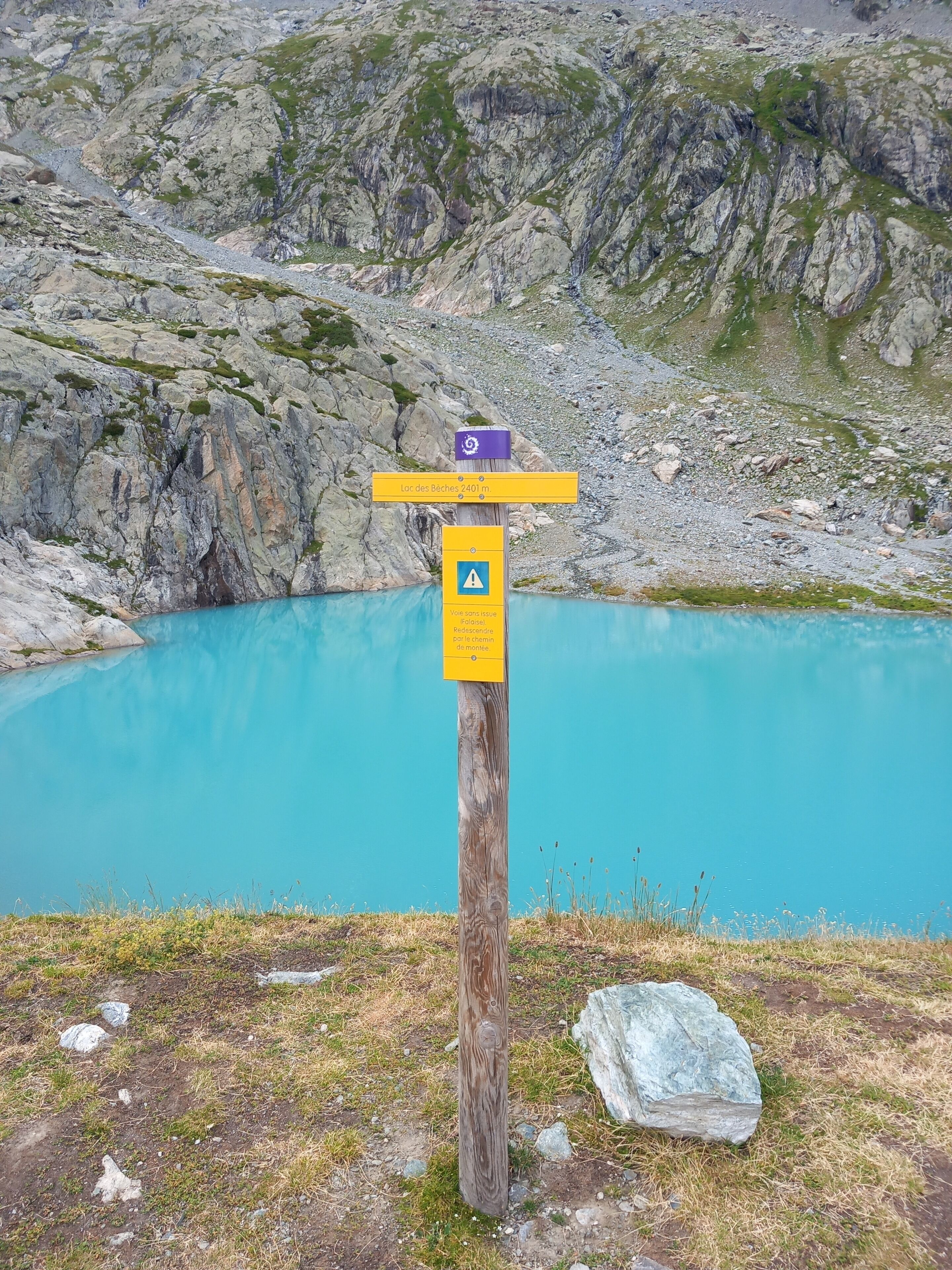 Un cartel de madera con indicaciones y un símbolo de advertencia, sobre un telón de fondo de una montaña rocosa y un lago turquesa.
