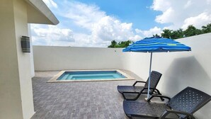 A rooftop pool area with a blue-striped umbrella, two lounge chairs, and a small hot tub.