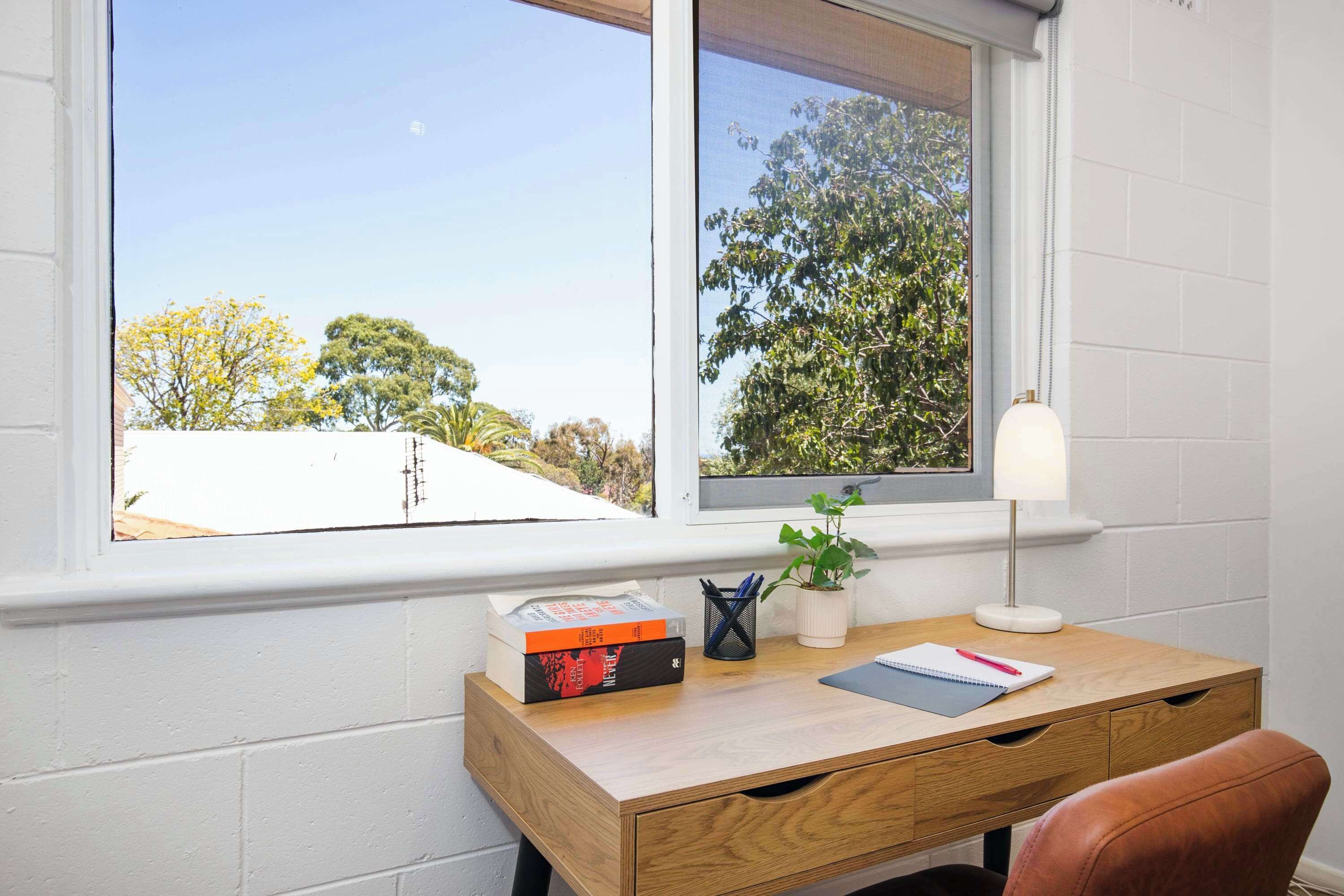 Un bureau avec une lampe, un carnet et une plante, près d’une fenêtre donnant sur des arbres.