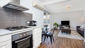 Eine moderne Küche mit Gasherd, Backofen und weißen Schränken. Es gibt einen Essbereich mit einem Glastisch und Stühlen sowie ein Wohnzimmer mit einer Couch und einem Fernseher.