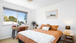 Ein Schlafzimmer mit Bett, Schreibtisch, Stuhl, Standventilator, Nachttischlampe und einem gerahmten Bild an der Wand.
