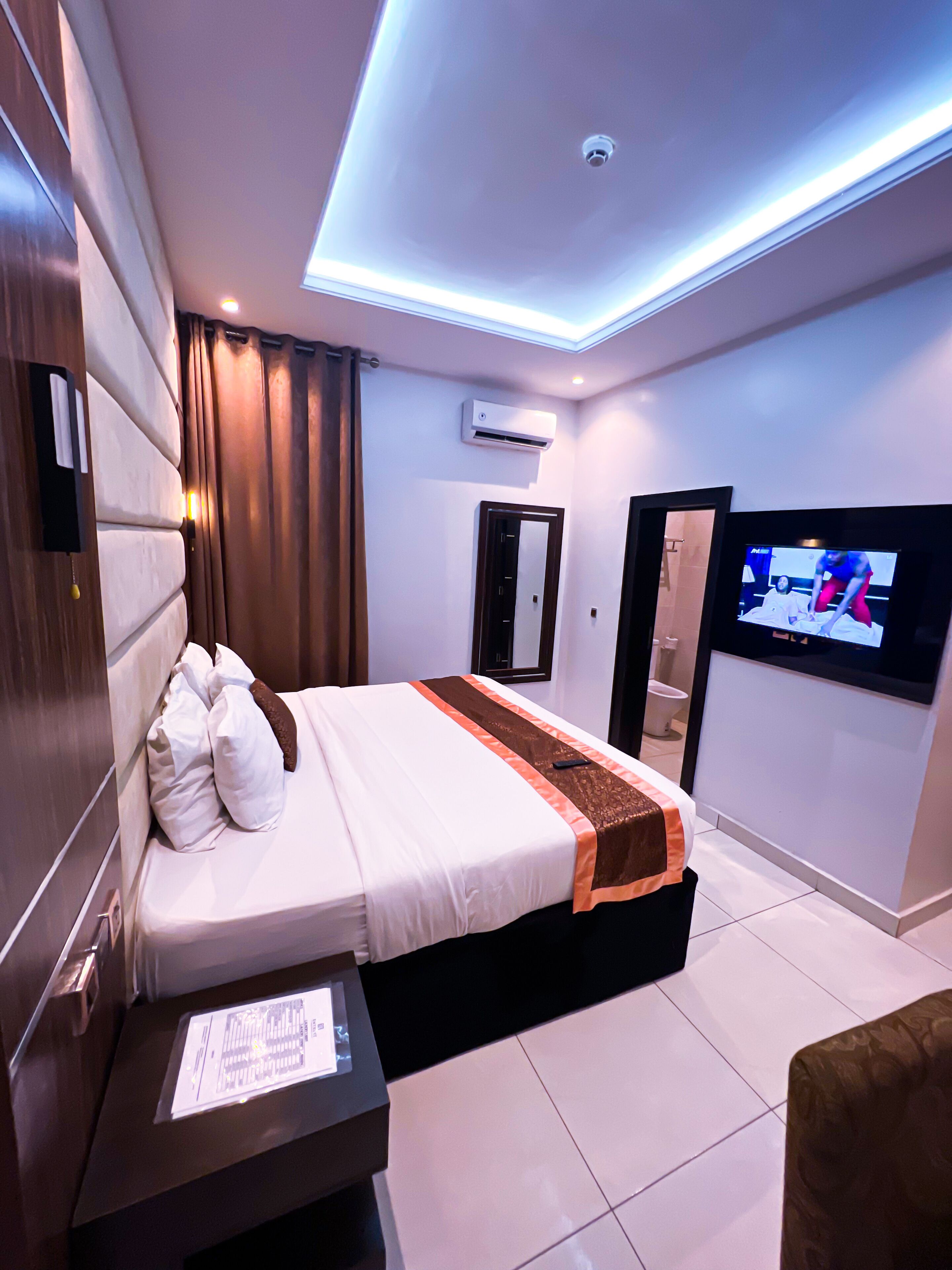 A modern hotel room with a large bed, a flat-screen TV, an air conditioning unit, and a bedside table with a remote control.