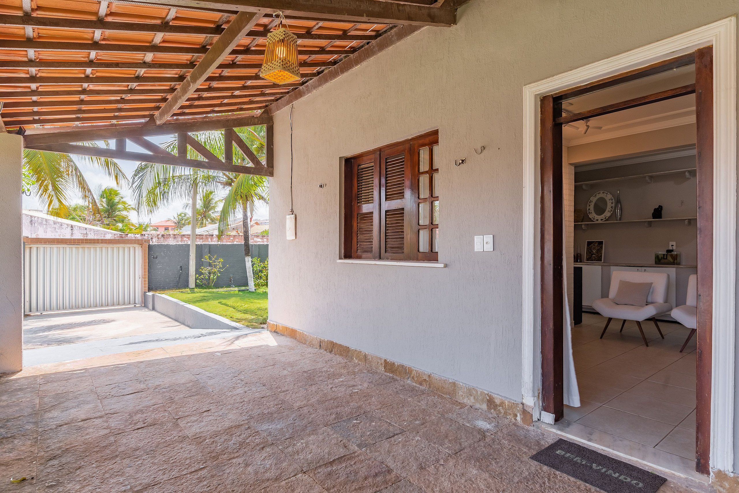 A covered outdoor area with a wooden ceiling, a window with shutters, and a door leading to an interior space with a white chair and shelves.