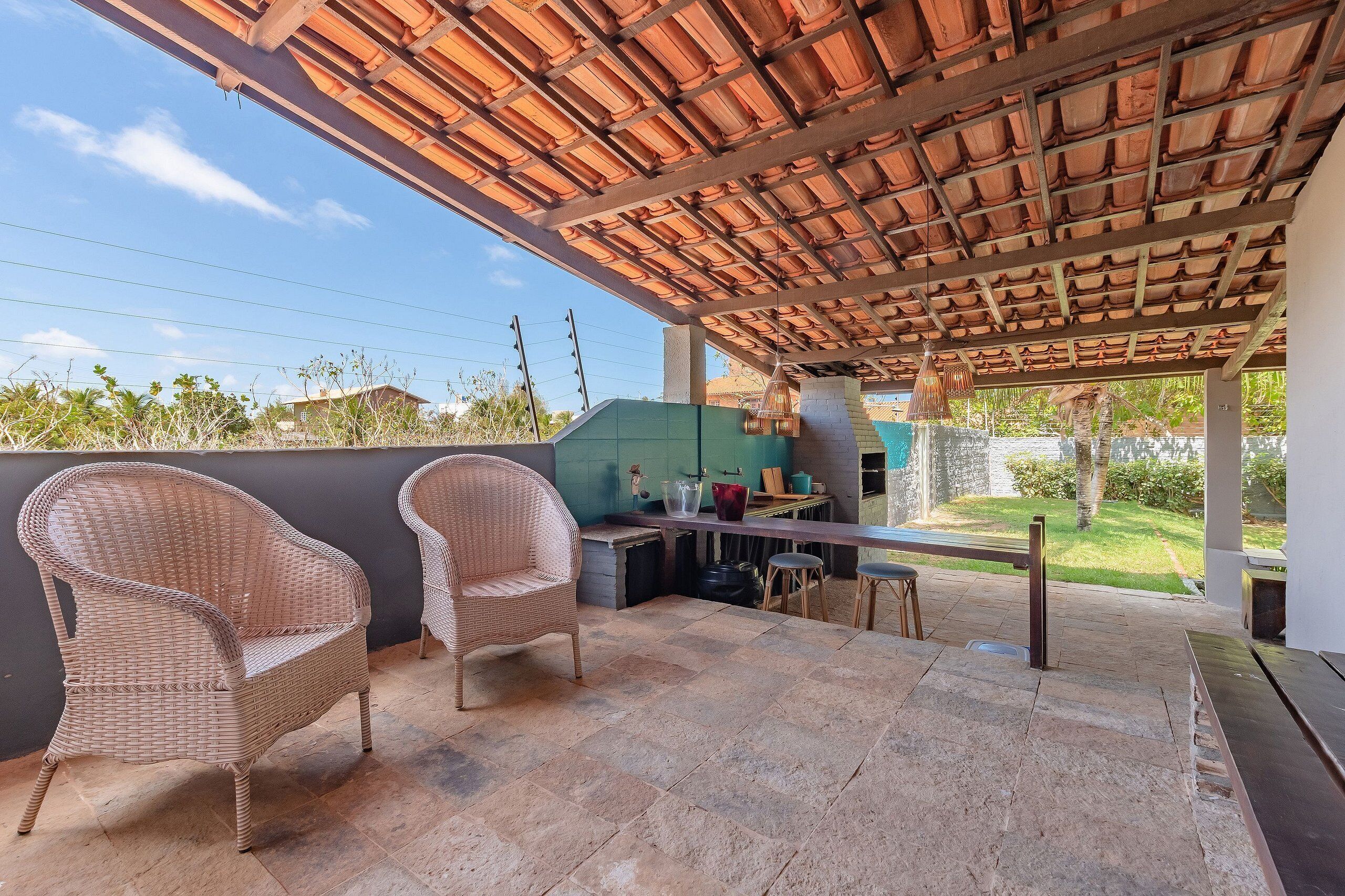 Ein überdachter Patio-Bereich mit Korbstühlen, Steinplattenboden, einer Küchenzeile mit Grill und Blick ins umliegende Grün.