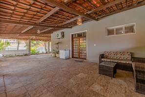 An outdoor covered patio area with a stone floor, a wooden ceiling, a white air conditioning unit, a wicker sofa, and a small table. - Casa melhor de Porto das D - Carpediem (Aquiraz)