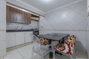 A kitchen with white cabinets, a wooden shelf, a tiled backsplash, a sink, and a dining table with colorful chairs. - Casa melhor de Porto das D - Carpediem (Aquiraz)