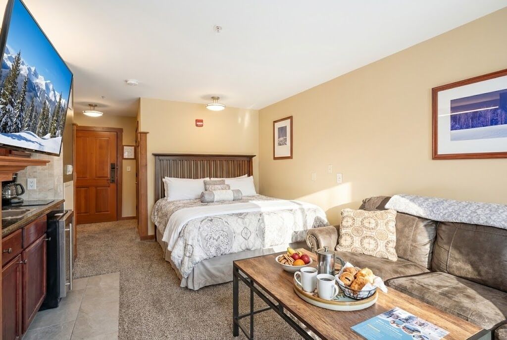Habitación de hotel con cama, sofá, mesa de comedor con desayuno, televisor y zona de cocina.