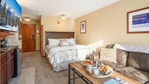 Ein Hotelzimmer mit Bett, einer Couch, einem Esstisch mit Frühstück, einem Fernseher und einer Küchenzeile.