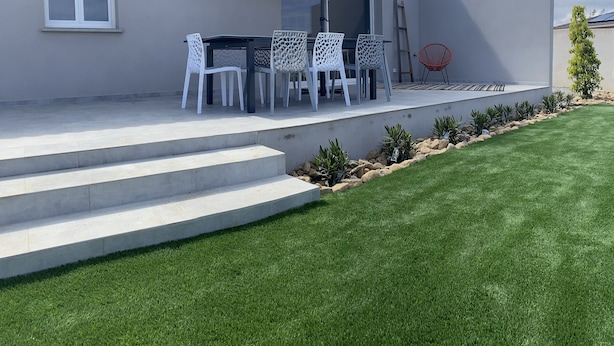 Un patio con una mesa y sillas, una sombrilla y un pequeño jardín con césped y plantas.