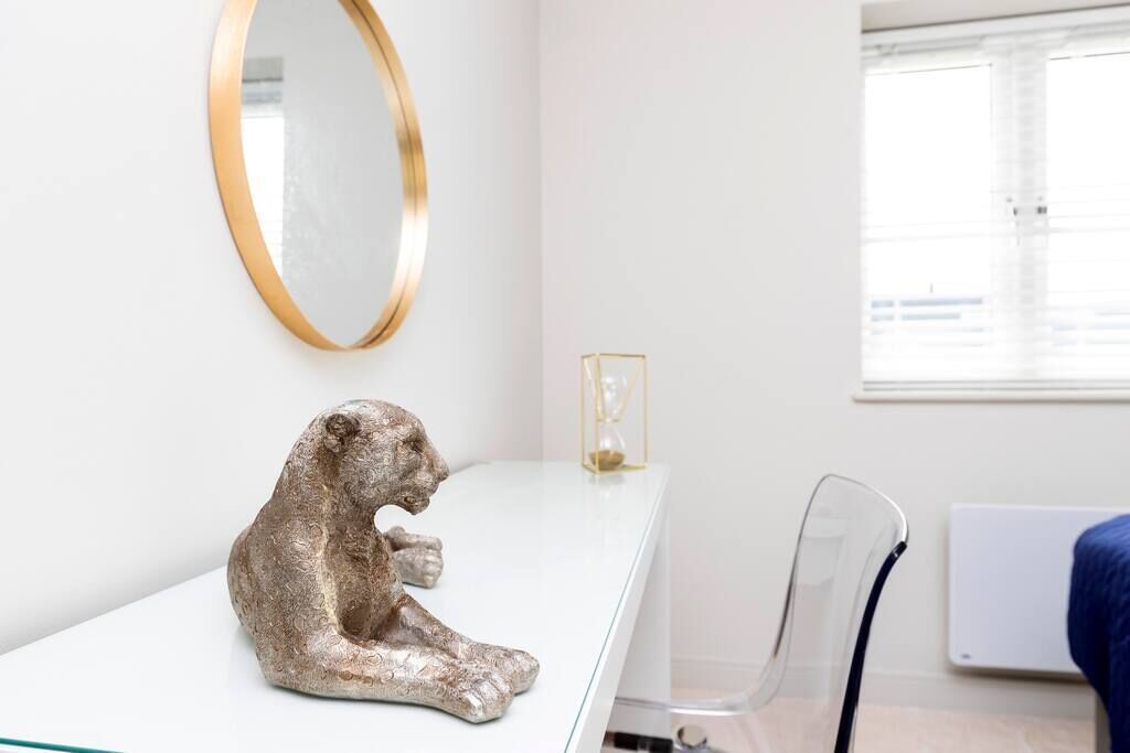 Une pièce au décor minimaliste, avec un bureau blanc, un miroir à cadre doré, une chaise transparente et une fenêtre équipée de stores.