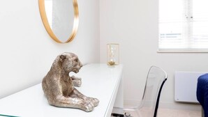 Une pièce au décor minimaliste, avec un bureau blanc, un miroir à cadre doré, une chaise transparente et une fenêtre équipée de stores.