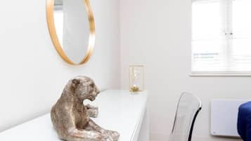 Une pièce au décor minimaliste, avec un bureau blanc, un miroir à cadre doré, une chaise transparente et une fenêtre équipée de stores.