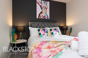 A modern bedroom with a tufted headboard, colorful pillows, a tie-dye blanket, and a large abstract painting on the wall.