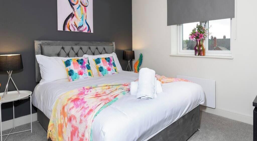Une chambre à coucher moderne avec une tête de lit rembourrée de couleur grise, des coussins colorés, un grand tableau abstrait et une fenêtre offrant une belle vue.