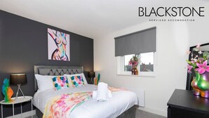 A modern bedroom with a grey upholstered headboard, a colorful bedspread, and a large abstract painting on the wall. - Avery House - 1 Bed with Outdoor Terrace (Darlington)