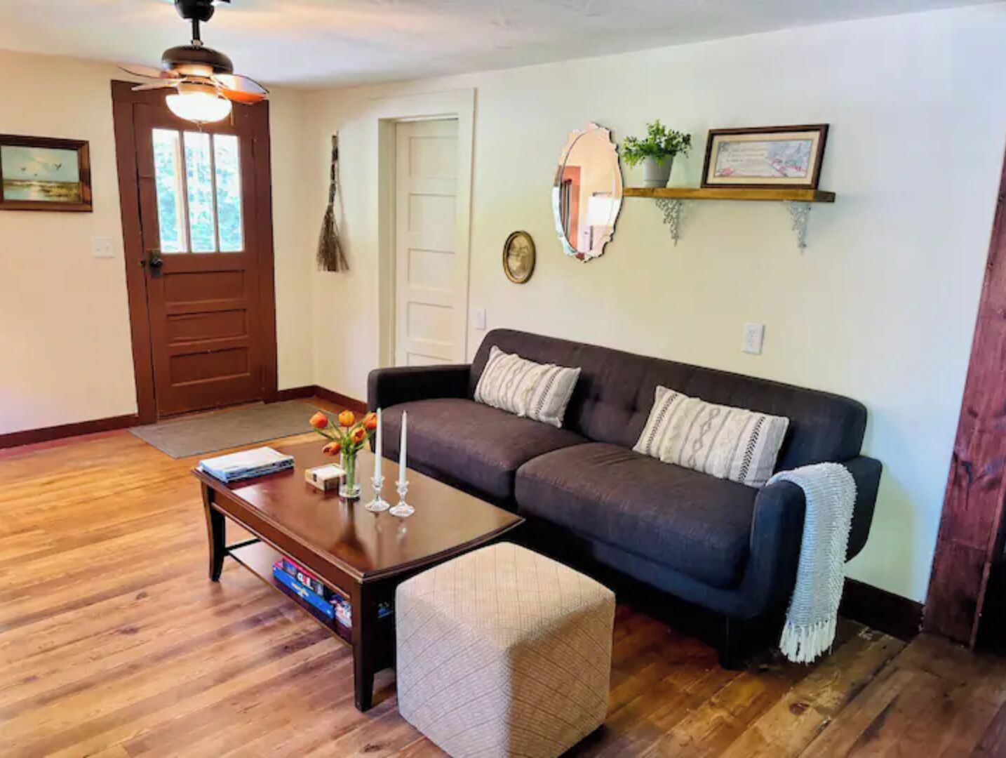 Un salon avec un canapé marron foncé, une table basse en bois et un petit pouf. On y trouve une porte surmontée d’un miroir et un ventilateur de plafond.