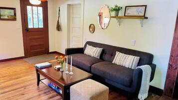 A living room with a dark brown sofa, a wooden coffee table, and a small ottoman. There is a door with a mirror above it and a ceiling fan.