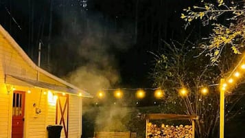 A rustic outdoor seating area with a fire pit, surrounded by red chairs, under string lights, with a stack of firewood and a building in the background.