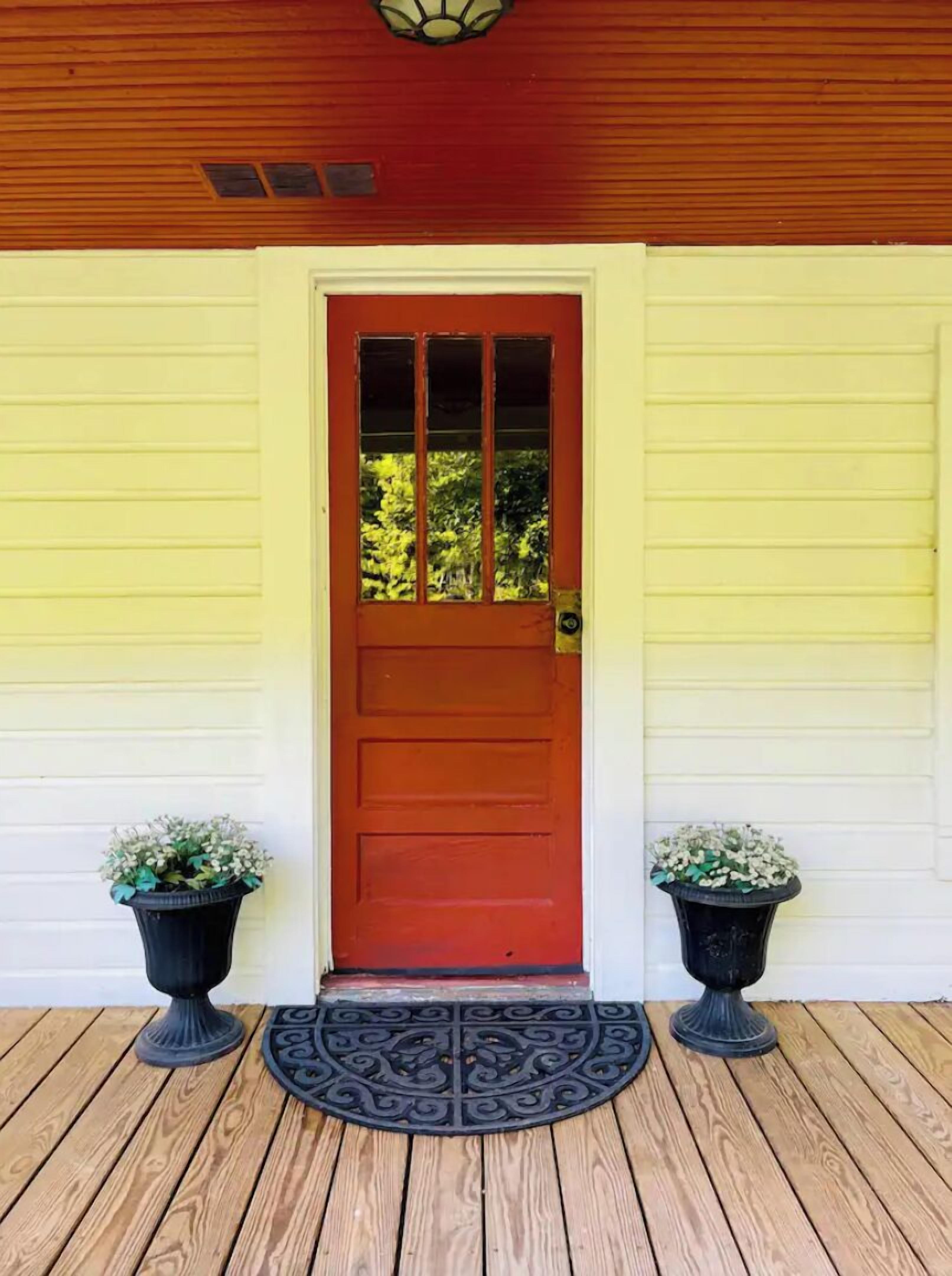 Une porte en bois, dotée d’un cadre rouge et de panneaux de verre, est encadrée de deux jardinières noires ornées de fleurs blanches, le tout posé sur une terrasse en bois sur laquelle se trouve un paillasson décoratif en métal.