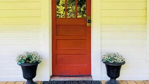 A wooden door with a red frame and glass panels, flanked by two black planters with white flowers, on a wooden deck with a decorative metal doormat.
