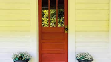 A wooden door with a red frame and glass panels, flanked by two black planters with white flowers, on a wooden deck with a decorative metal doormat.