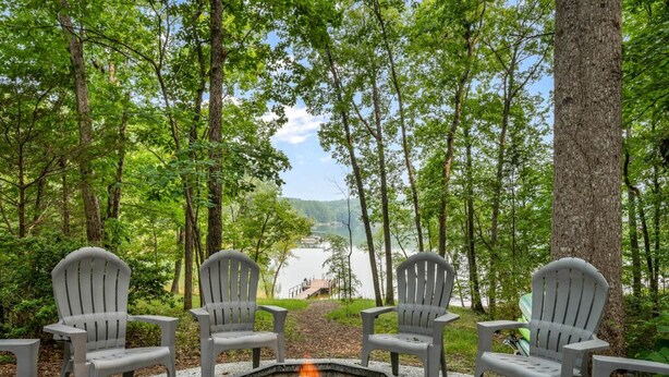 Un foyer est entouré de chaises Adirondack, et l’ensemble est installé face à un lac, avec un quai et des montagnes en arrière-plan.