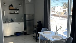 Une cuisine moderne avec des armoires blanches, un four encastré et une machine à café. On y trouve un coin repas avec une table blanche et deux chaises, ainsi qu’une grande fenêtre offrant une vue sur un paysage enneigé.