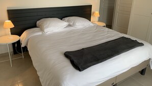 A neatly made bed with white linens and a dark blanket, flanked by two bedside tables with lamps, in a room with a plain wall and a door.