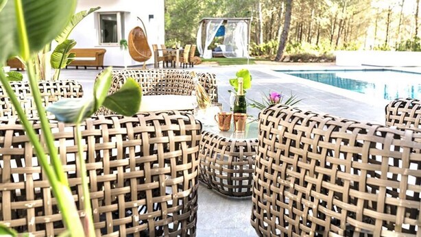 Wicker furniture arranged around a pool area with a white building in the background. - Charming Villa in the Nature of Santa Gertrudis (Islas Baleares)