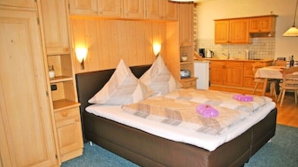 A bedroom with a large bed, wooden cabinets, and a kitchenette in the background. - Vacation apartment I - Guesthouse Kamml (Inzell)