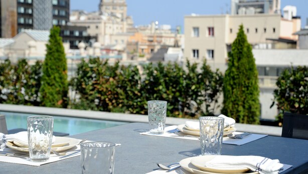 A rooftop dining area with a pool view, tables set with plates and glasses, and a cityscape in the background.